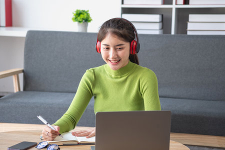 Asian woman with a beautiful face in a green long-sleeve shirt sitting and relaxing watching movies Listening to music on a laptop, mobile phone, and headphones happily inside living room at home.の写真素材