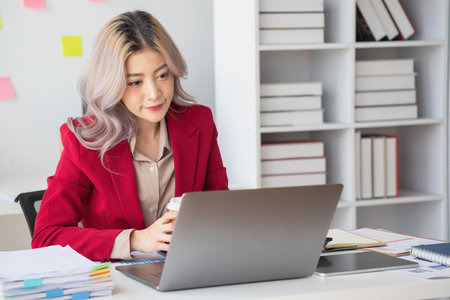 A businesswoman in a red suit holding a cup of coffee to relax in a modern office room before calculating business profit for the company.の写真素材