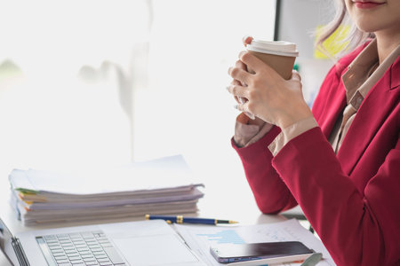 A businesswoman in a red suit holding a cup of coffee to relax in a modern office room before calculating business profit for the company.の写真素材