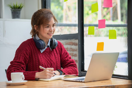 Beautiful Asian businesswoman sitting at a table with computer wearing headphones with microphone Looking at the laptop camera Happy businesswoman educating customers online.の写真素材
