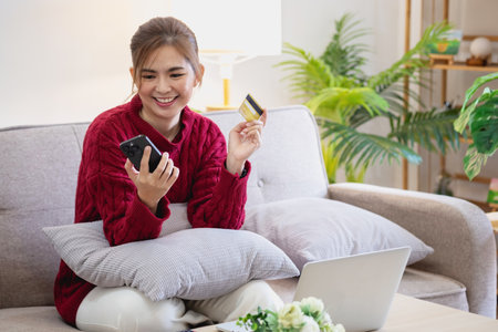 A young Asian woman with a happy smile holds a credit card and uses a smartphone to shop online Online payment concept.の写真素材
