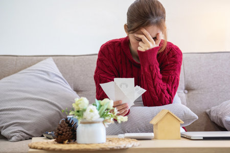 A young Asian woman sits on a sofa in her home, feeling worried and frustrated about her monthly expenses. Various utility bills.の写真素材