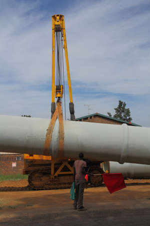 A worker waves a red flag as a warning signal while a large pipe is lifted off a truck