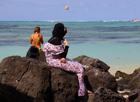 Ile aux Cerfs, Mauritius - unidentified young woman in traditional Muslim dress looks out over the ocean as another unidentified bather in a bikini stands nearbyのeditorial素材