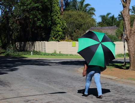 An unidentified African woman uses a colorful umbrella to protect herself against the harsh African sun on her walk home, image in landscape formatの写真素材