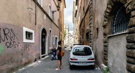 Rome, Lazio, Italy. July 25, 2017: Narrow cobblestone street in the center of town with a young couple. The man is on his knees taking a picture,のeditorial素材