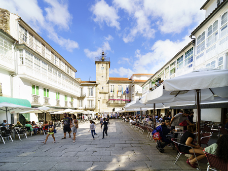 Pontedeume, Galicia / Spain. July 29, 2017. Town square with stone block floor, with bar terraces with people drinking and children playing. In the background the tower of the town hallのeditorial素材