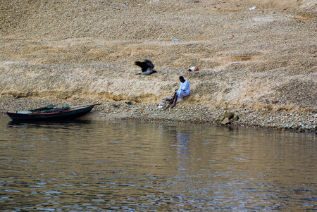 Nile river, near Aswan, Egypt, February 21, 2017: Egyptian fisherman dressed in the typical white djellaba resting on the side of his boat stranded on the shoreのeditorial素材