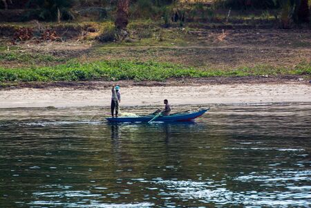 Nile river, near Aswan, February 16, 2017: Small blue fishing boat typical of the river with two fishermen, one of them standing by handling the net and the other paddlingのeditorial素材