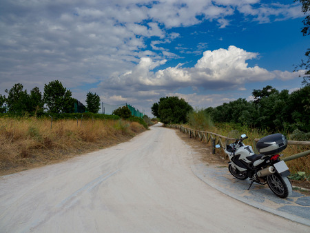 Dirt road with dry yellow vegetation and green trees, with a motorcycle parked at the beginning of the roadの写真素材