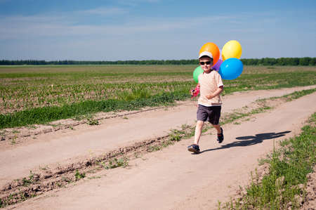 Little brave boy with colorful balloons running by the country roadの写真素材