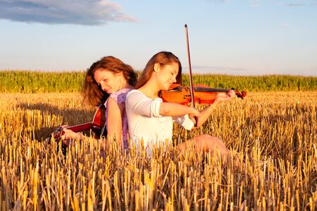 Two young women playing guitar and violin outdoors on the field in summer evening sitting back to backの写真素材