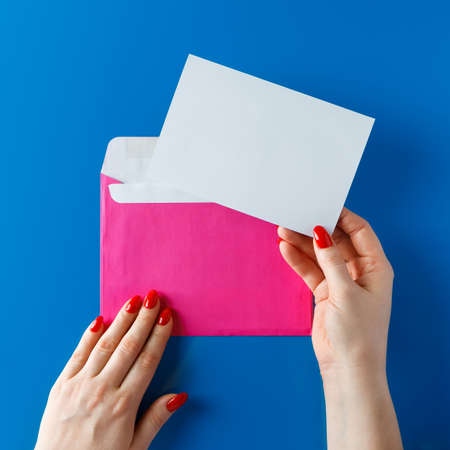 Pink envelope with a blank card in hands on a blue background. Pink envelope with a greeting card in the hands of a young woman.の写真素材