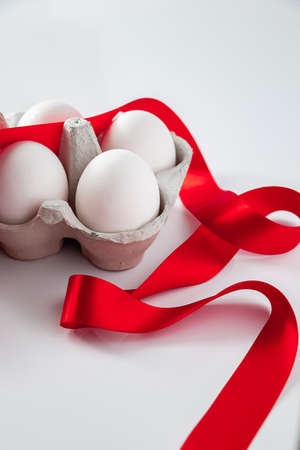 Easter eggs in a cardboard box with a red festive ribbon on a white background. White easter eggs in...