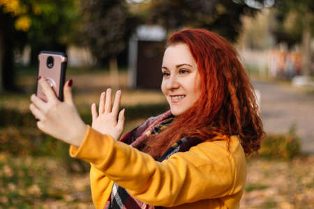 Young red-haired woman is video chatting. She uses modern smartphone. Lady smiles and walks in park...