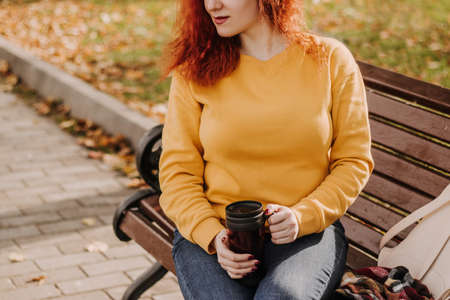 Portrait of young red-haired woman sitting in park on bench with mug of coffee. Lady is wearing yellow sweatshirt and holding reusable cup. Lifestyle on a sunny autumn day.の写真素材