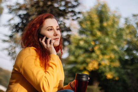 Young red-haired woman in yellow sweatshirt talking on phone. Lady walking in autumn park on a sunny...