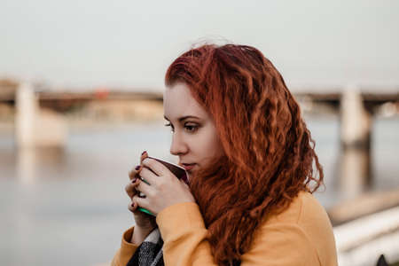 Portrait of young red-haired woman against background of city river and bridge. She drinks coffee from paper cup and enjoys autumn view. Copy space.の写真素材