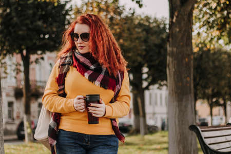 Portrait of young red-haired woman in yellow sweater and sunglasses. She holds reusable coffee mug in her hands and walks through park on sunny autumn day. Eco-friendly lifestyle.の写真素材