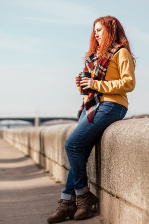 Young woman in yellow sweater sits on embankment and drinks coffee from reusable mug. She is enjoying autumn sunny day. Eco-friendly lifestyle, relaxation.の写真素材