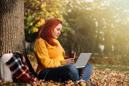 Young woman drinks coffee and works using laptop in nature. Remote work on the Internet. Rest and...