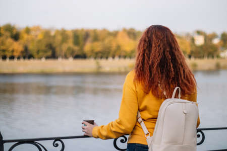 Back view of red-haired woman. Lady with white backpack enjoys autumn view and drinks coffee from paper cup. Eco-friendly lifestyle, copy space.の写真素材