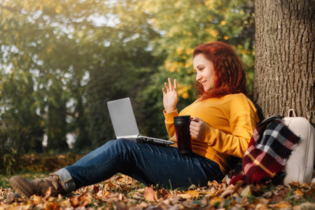 Red-haired woman in yellow sweater is video chatting. She uses laptop to communicate and work in Internet. Walk on autumn sunny day.の写真素材