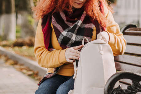 Young red-haired woman in yellow sweater sits on bench and looks for something in her bag. Walk on a sunny autumn day.の写真素材