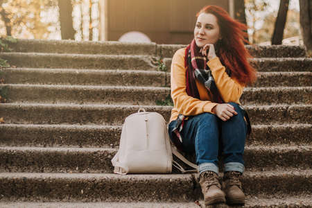 Young red-haired woman sits on stairs in park. Lady in yellow sweater is enjoying sunny day. Urban tourism. Lifestyle, copy space.の写真素材