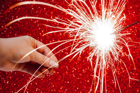 New Year celebration. Womans hand holds bright sparkler on red background. Christmas night. Bright sparks of fire and bokeh. Selective focusの写真素材