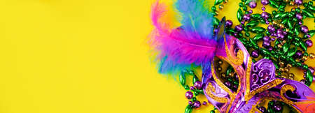Carnival mask with feathers on yellow background. Multicolored beads in green, gold and purple. Mardi Gras or Fat Tuesday symbol. Copy space, banner format.の写真素材