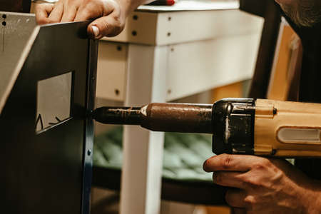 Male worker uses electric riveting gun. Hand holding riveting machine and placing rivets. Working workshop of carpenter or mechanic.の写真素材
