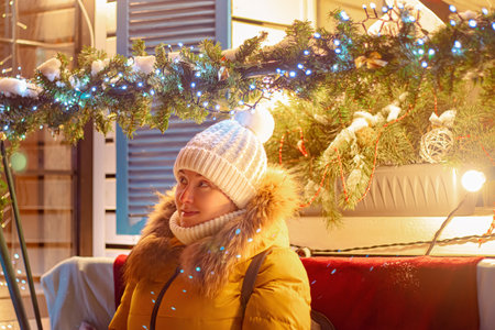 Young woman tourist in knitted hat looks at festive Christmas city decorations. Christmas market, night lights.の写真素材