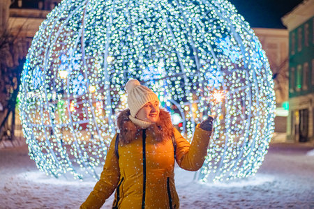 Young woman in knitted hat holds sparkler in her hands. Night festive street, shiny city decorations for Christmas.の写真素材