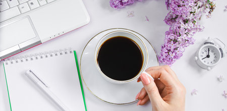 Female hands holding cup of espresso coffee. Alarm clock and lilac flowers on light background. Spring flatlay. Morning breakfast in spring. Banner format.の写真素材