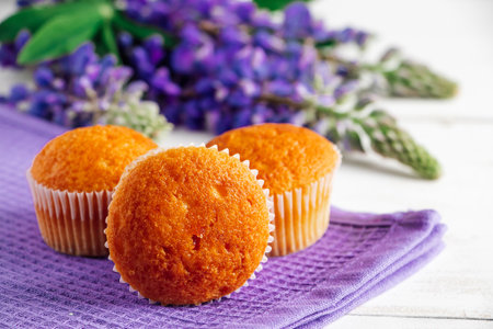 Soft fresh muffins on textile napkin. Delicious pastries and purple lupine flowers on white wooden background. Preparing dessert in confectionery.の写真素材