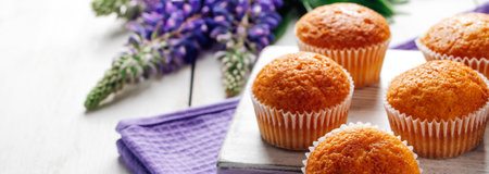 Soft fresh muffins on textile napkin. Delicious pastries and purple lupine flowers on white wooden background. Preparing dessert in confectionery. Banner format.の写真素材