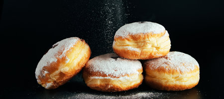 Powdered sugar is poured onto donuts. Traditional Jewish dessert Sufganiyot on black background. Celebrating Judaism holiday. Cooking fried Berliners. Banner formatの写真素材