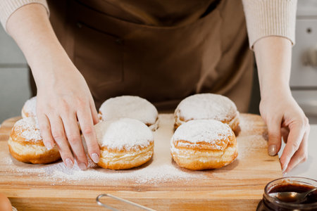Woman prepares fresh donuts with jam in home kitchen. Cooking traditional Jewish Hanukkah sufganiyot. Hands sprinkle Berliners with powdered sugar.の写真素材