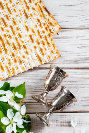 Happy Passover flatlay. Jewish matzo bread and wine glasses on white wooden background. Holiday greeting card with flowers. Copy space. Vertical photo.の写真素材