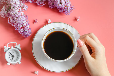 Female hands holding cup of espresso coffee. Alarm clock and lilac flowers on light background. Spring flatley. Morning breakfast in spring.の写真素材
