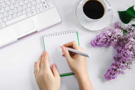 Spring flatlay. Woman works at home on laptop in morning. Cup of coffee and lilac flowers on table. Women's hands write in notepad.の写真素材
