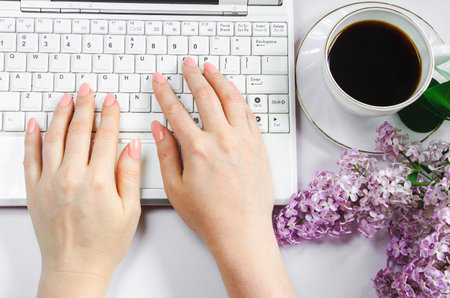 Spring flatlay. Woman works at home on laptop in morning. Cup of coffee and lilac flowers on table. Women's hands write in notepad.の写真素材