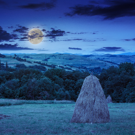 a pair of haystacks and a tree on a green meadow at the foot of the mountain at night in moonlightの写真素材