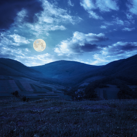 Stack of hay on a green meadow in the mountains in the morning under a blue summer sky at night in full moon lightの写真素材