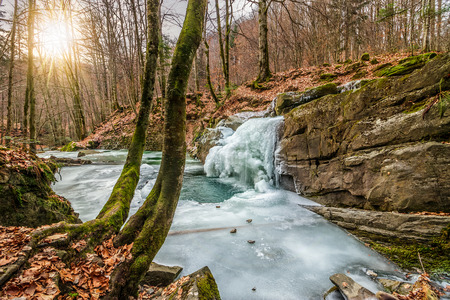 frozen waterfall over the huge boulder on the river among empty forest with old brown foliage on the groundの写真素材
