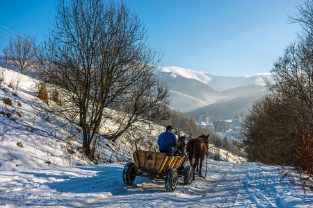 man riding cart with twoo horses down the hill to village in snowy mountainsの写真素材