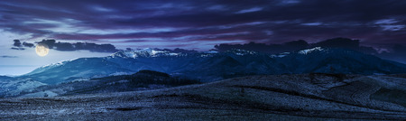 Early spring highland landscape. Panorama of rural fields on hill side in mountains with snowy peaks at night in full moon lightの写真素材