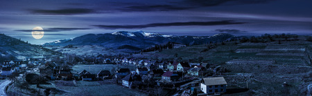 Early spring rural landscape. Panorama of village near the fields on hill side at the foot of the  mountains with snowy peaks at night in full moon lightの写真素材
