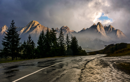 Travel destination concept image. Composite landscape of High Tatra mountain ridge. Curve asphalt road through spruce forest. Peaks lit by the sun in stormy weather with dramatic sky.の写真素材